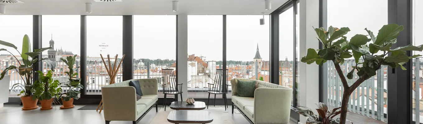 Top-floor lounge with sofas, plants and floor-to-ceiling windows overlooking the rooftops of central Prague